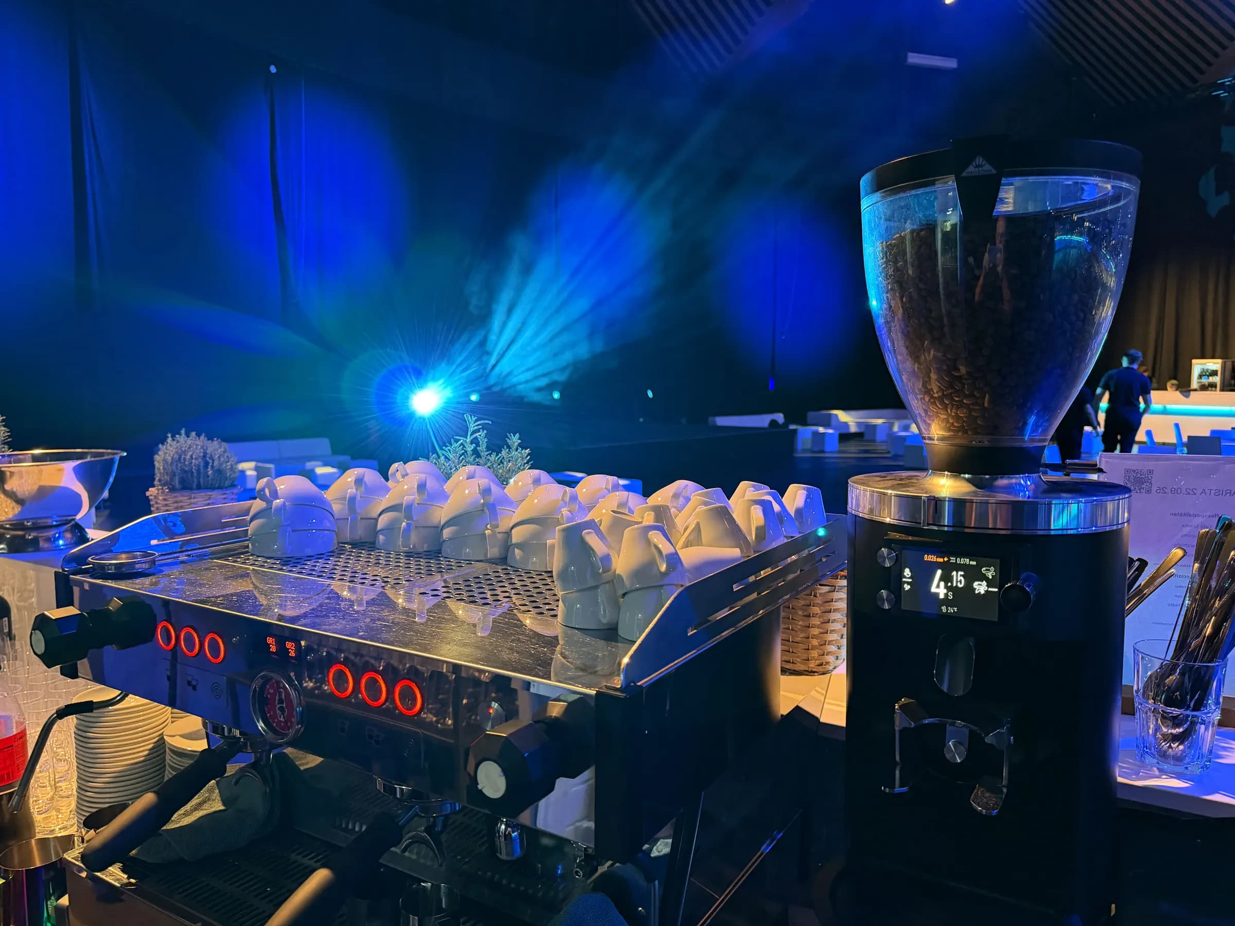 Zweigruppige Espressomaschine mit vorgewärmten Tassen und Mühle auf einer Eventbar, im Hintergrund blaues Bühnenlicht