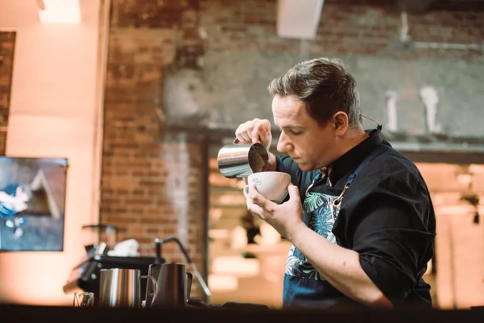 Barista gießt Milch aus einem Edelstahlkännchen in eine Tasse